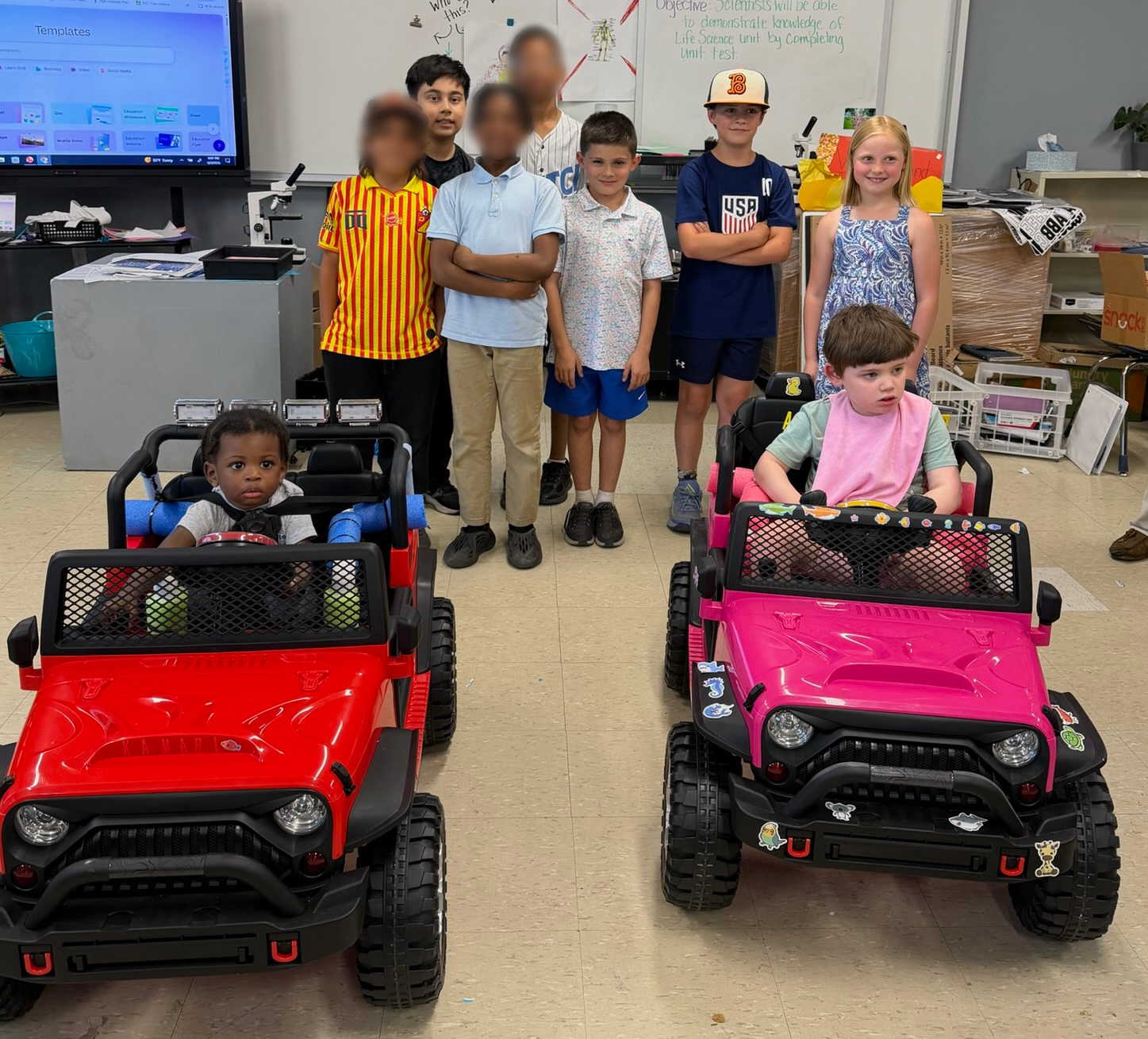 Students posing with two custom-built adaptive ride-on cars in a classroom