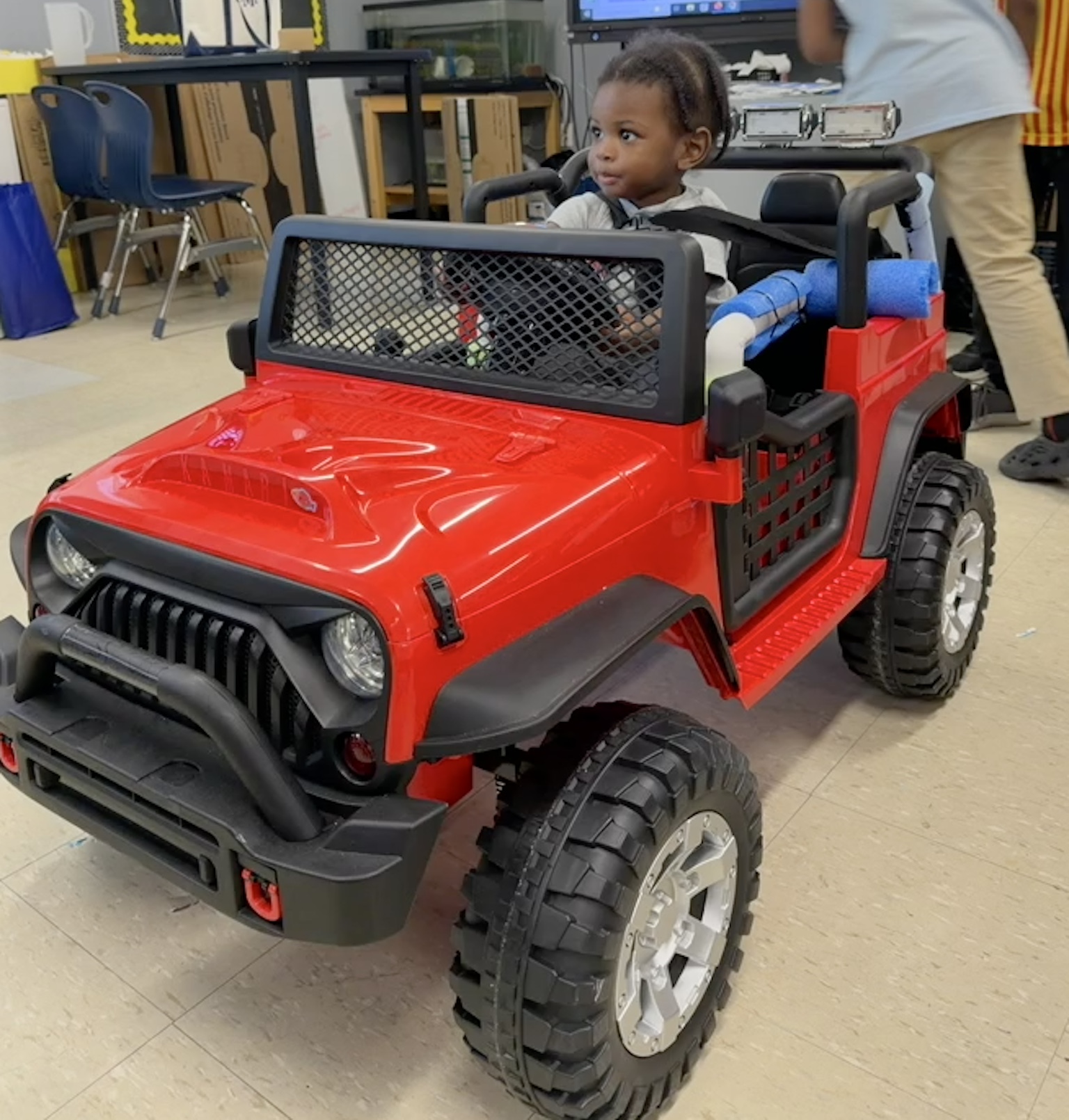 A young child smiling in a colorful adaptive ride-on car built by students