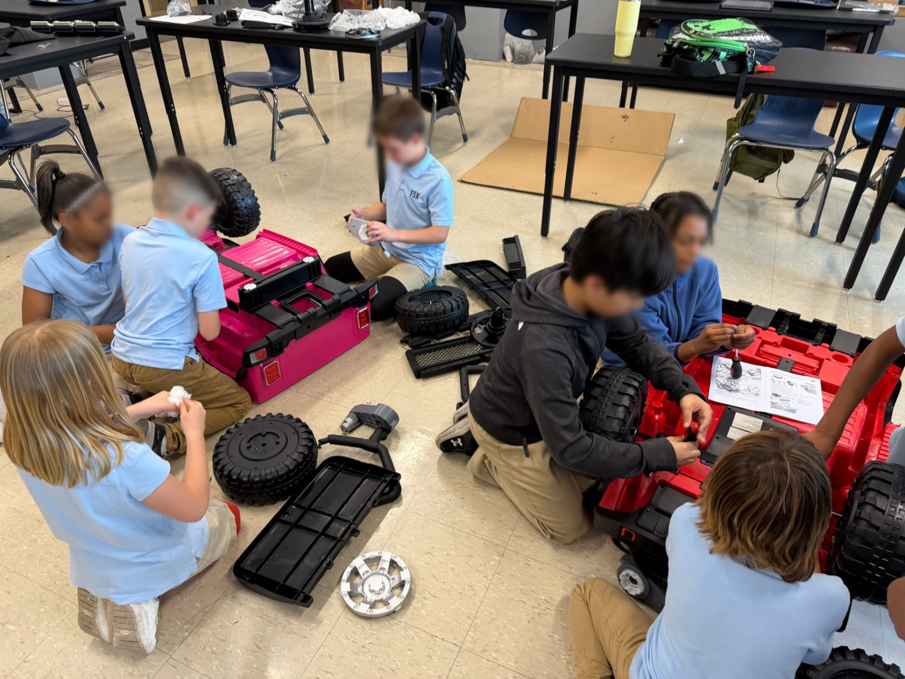 Elementary students working on the floor to assemble pink and red ride-on cars