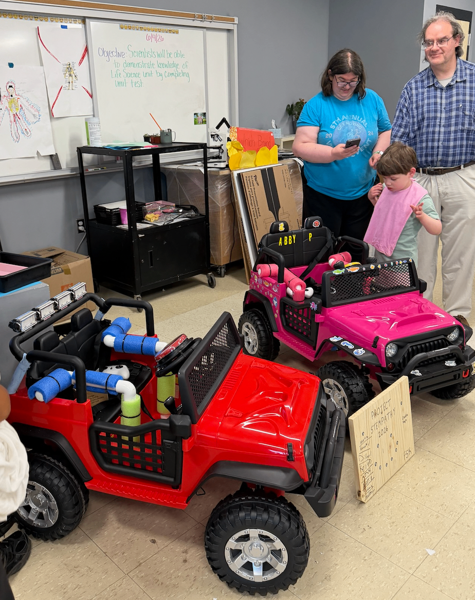 Two completed adaptive ride-on cars, red and pink, with a family in the classroom