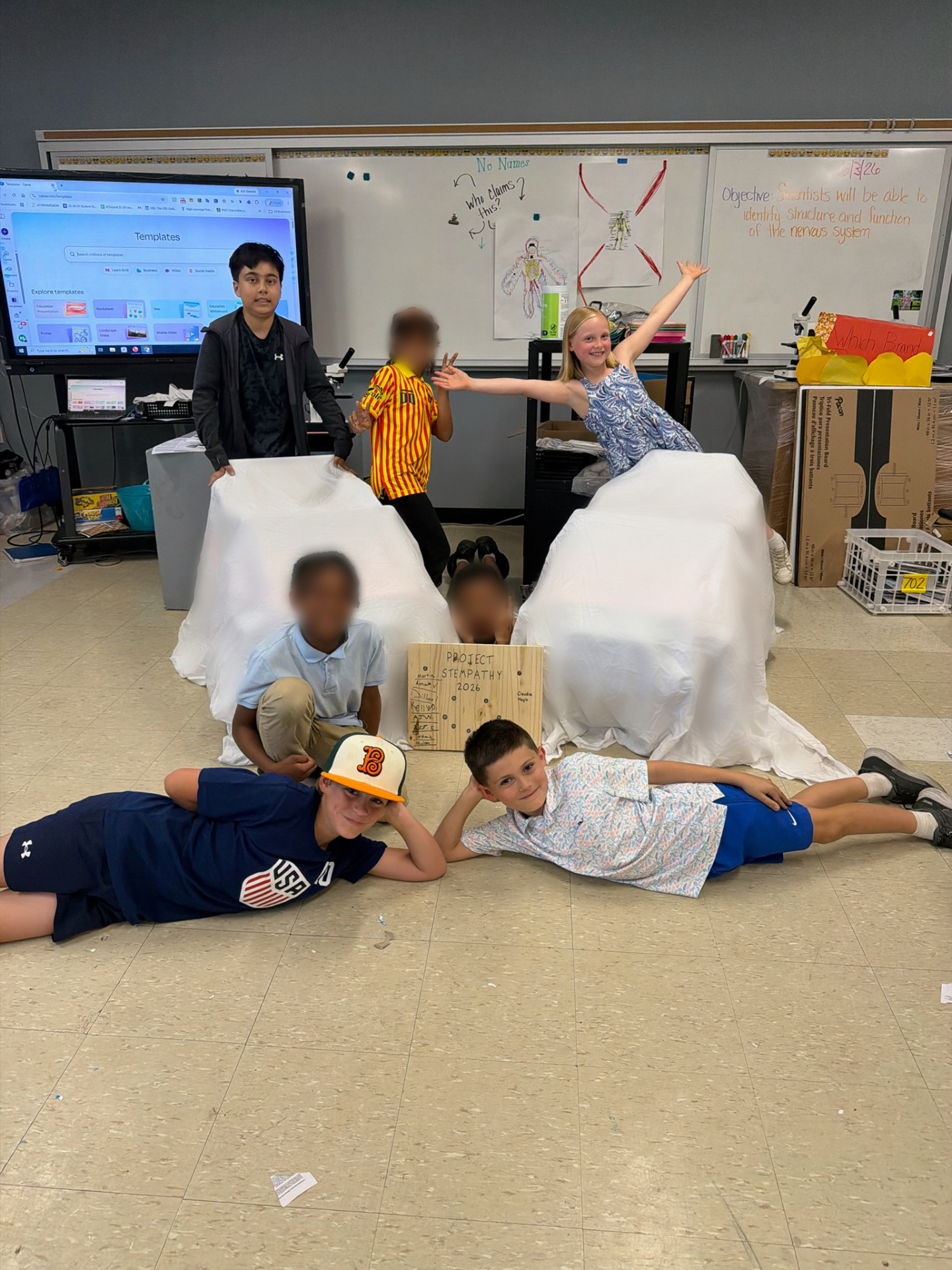 Students posing playfully with two ride-on cars covered by sheets for a big reveal