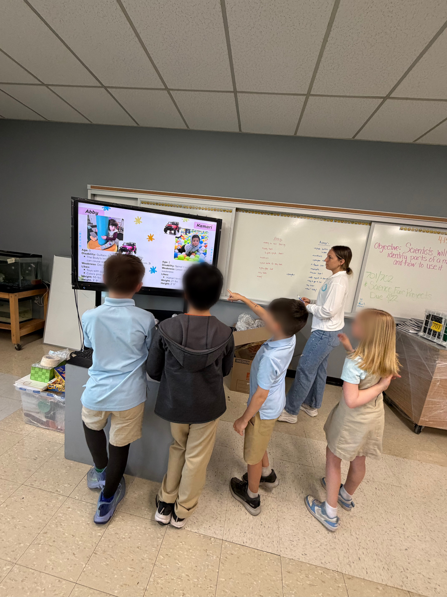 Students gathered around a smartboard learning about the children who will receive adaptive cars