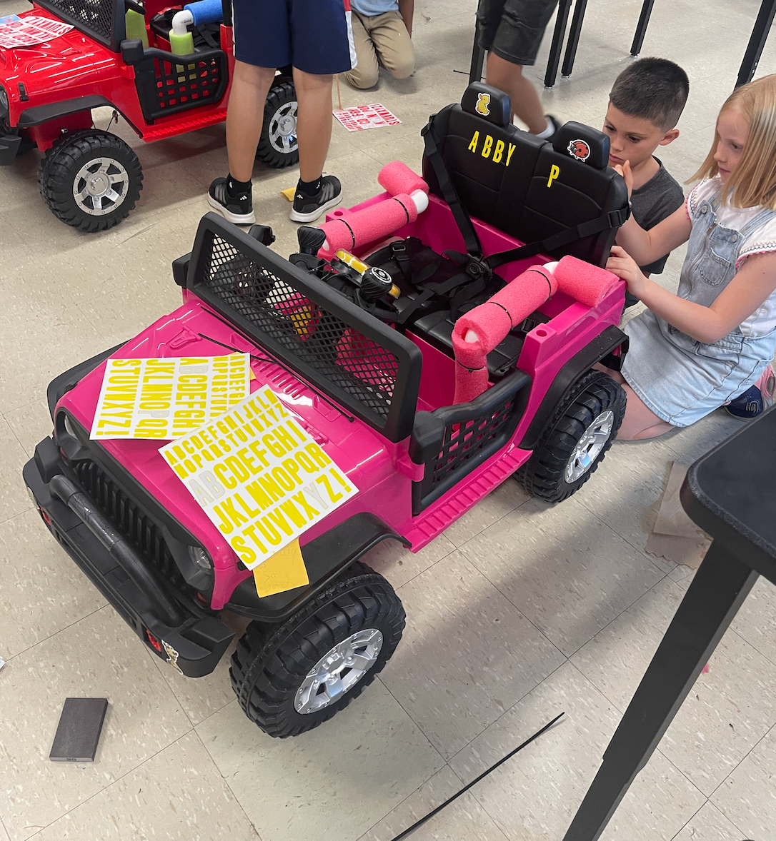 Students decorating a pink adaptive Jeep with colorful alphabet stickers