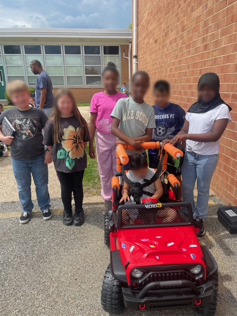 Students gathered around a child seated in a finished red adaptive ride-on car outside the school