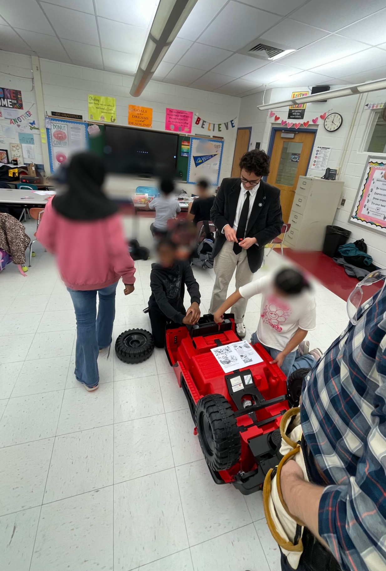 Students assembling a red ride-on car while a volunteer helps in the classroom